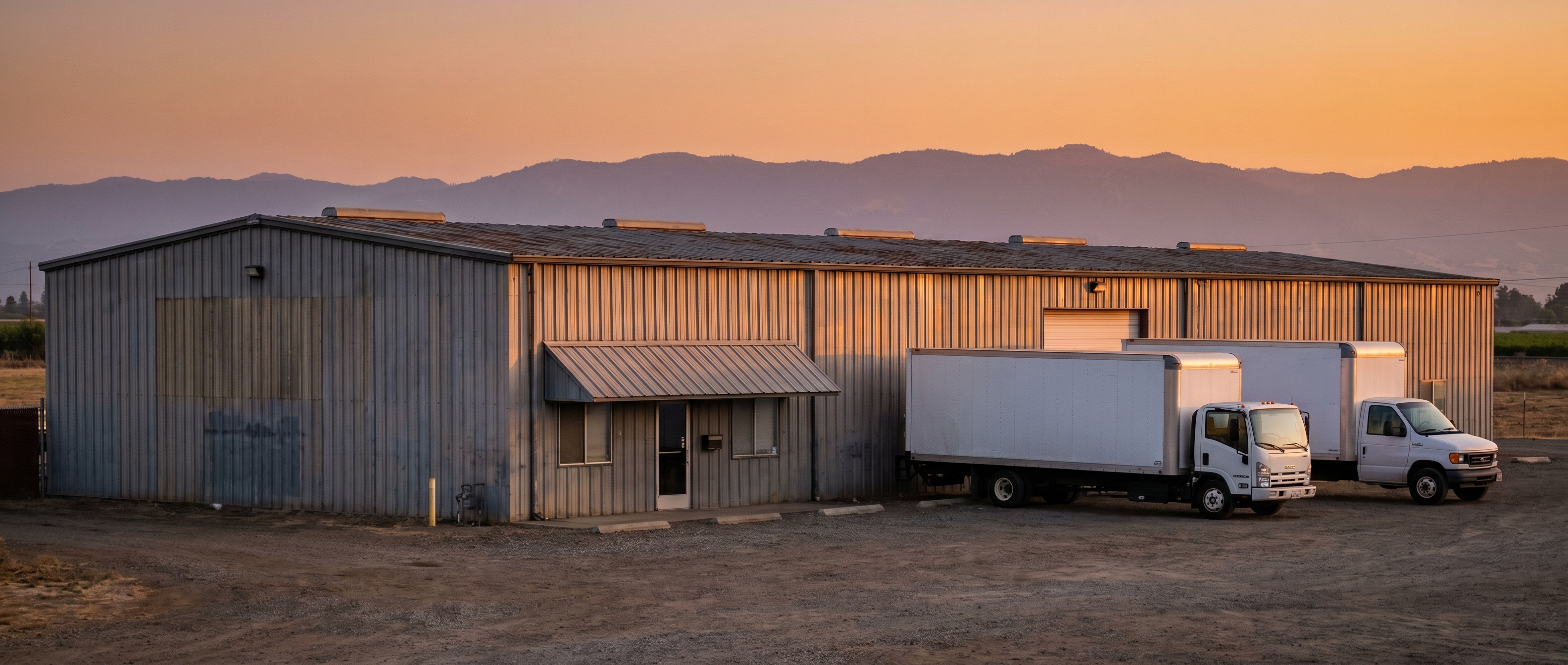 Helios warehouse exterior at golden hour, three white box trucks parked, metal-clad industrial building.
